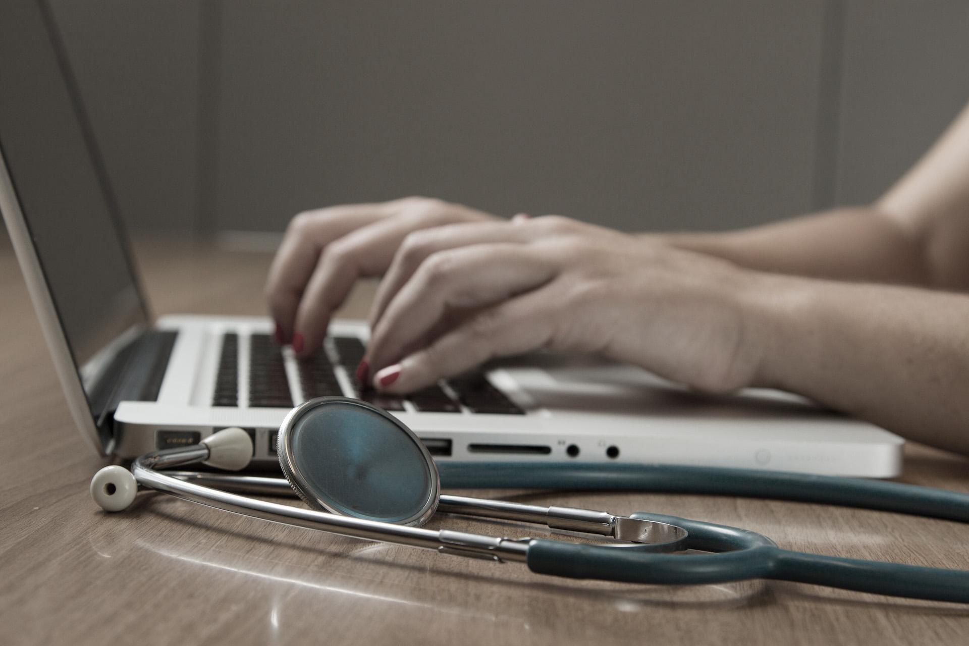 Hands type at a laptop while a stethoscope lays beside